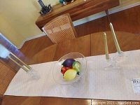 Overall view of two clear glass candle holders with gold candles and a glass fruit bowl filled with colorful glass fruit on a table.