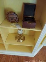 Shelf with wooden spiral wall clock and wooden box clock side by side, above brass anniversary clock with glass dome on lower shelf.
