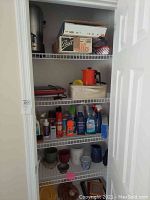 Closet shelving with assorted kitchenware, cleaning supplies, mugs, jars, tins, and small appliances such as electric griddle and coffee pot visible.