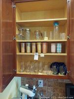 Opened wooden cabinet showing three shelves containing assorted Mason jars, glasses, tumblers, and glass mugs.