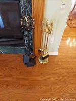 Two sets of fireplace tools placed side by side on a wooden floor, one black with brass handles and one all brass finish.