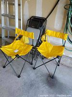 Two yellow folding camp chairs fully extended side by side with accompanying carry bags hanging from them, black metal frames and yellow vinyl seats and backs.