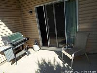 Wide shot of the Weber grill and metal chair positioned outside on concrete near a sliding glass door.