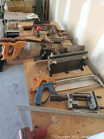 Photo shows multiple hand tools including two saws, a miter box, clamps, and a square ruler laid out on a wooden table.