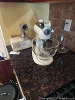 Full view of off-white KitchenAid stand mixer with attached glass mixing bowl and cylindrical metal grinder unit on counter, unplugged.