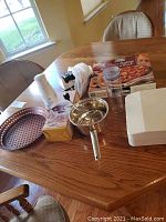 Full view on wooden table showing Zyliss chopper, pizza pan, Williams Sonoma chopper box, Chef's Choice electric sharpener, food mill, pizza stone and rack, scale, herb container, and kitchen utensils.