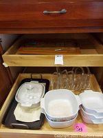 Drawer containing a black rectangular griddle pan with handles, stacked CorningWare white ceramic baking dishes with floral decoration, and clear glass lids.