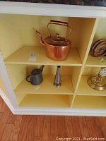 Shelf display of copper tea kettle along with metal pitcher and metal funnel placed below it.