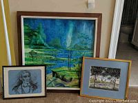 Three framed artworks stacked on floor against wall showing a large landscape painting, a smaller portrait sketch, and a medium cityscape landscape