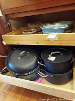 Wooden kitchen cabinet drawers open showing 8 frying pans and 2 pots stacked inside with lids