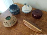 Five pottery pieces including a dark blue vase, cream double vase, brown trinket box, beige carved trinket box, and wooden coaster holder on wood surface.