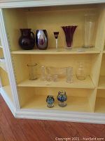 Wide view of shelf with multiple glass items including purple vases, candle holders, and clear glass pieces