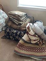 View of folded quilts and bed linens stacked on a black floral cushion/pillow on carpet near window.