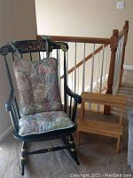 Rocking chair with floral cushions and wooden step stool positioned beside a staircase railing inside a house.