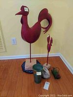 Full view of lot with two red metal rooster sculptures on stands, a galvanized metal watering can, a rabbit statue, and a hummingbird feeder on floor.