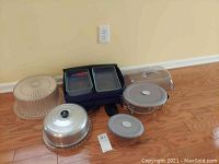 Full lot showing cake plates with covers, serving bowls, and carry case with food containers on wood floor.