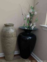 Two large decorative floor vases placed near a wall corner; one is tall beige with ribbed texture, the other is glossy black with floral stems