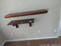 Two wooden wall shelves mounted on a white wall, one lifts off, the other screwed into the wall. Both have a brown finish and show wear.