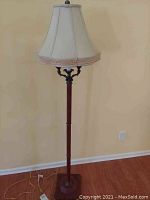 Full view of a tall wooden floor lamp standing on a hardwood floor against a light wall, showing its full height and structure including the base and the lampshade.