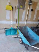 View of wheelbarrow, yellow dust pan, blue snow shovel and brooms in garage space