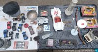 Wide overhead shot showing all items included in the lot including Speed Racer car, toy cars, camping lantern, police costume cap, electric fan, Leaning Tower of Pisa statue, door knobs, flexible mirror, and other small collectibles.
