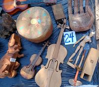 Overview of varied wooden musical instruments including carved violins, percussion instruments, hand drum, wooden dinosaur on wheels, wooden hand sculpture, artist doll, and tomahawk toy.