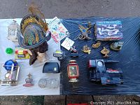 Full overview of all lot items placed on black plastic sheet. Items include air compressor, model car kit, brass figurines, miniatures, glass bottles, and clocks.