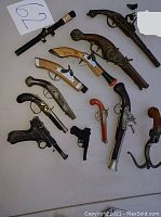 Overhead view of rifle scope and assortment of replica pistols on white surface