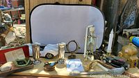 Photo showing the full spread of assorted items including the Norelco electric shaver box, miniature car models, skeleton figurine, gyroscope, couple of silver-tone cups, sundial, miniature coffee grinder, miniature bellows, note holder, large spoon and others all set on a display board outdoors.