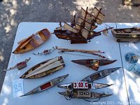 Collection of model boats and ships including wooden row boat, pontoons, kayaks, and ships in bottles arranged on white table