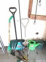 Garden tools including shovels, forks, watering cans, and hand tools arranged against a wall showing the shape and general condition.