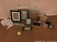 Photo showing a variety of picture frames in different styles and sizes arranged on carpeted floor against wall.
