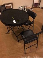 Photo showing round black textured folding table and three black padded folding chairs arranged around it on a beige carpet.