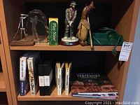 Two shelves with golf and baseball-themed books, figurines, and VHS tapes. Visible items include metal and acrylic golf figurines, golf-related books and memorabilia, wooden duck golf club cover, and baseball books and VHS tapes related to Brooklyn Dodgers and World Series.