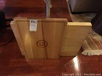 Two wood cutting boards side by side on wooden floor; the board in front has a juice groove and emblem, the other is plain wood.