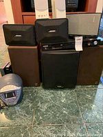 Photo showing two Bose bookshelf speakers, two KEF floor-standing speakers, Samsung DVD player, second DVD player, and an unknown black subwoofer or speaker on floor tiles.