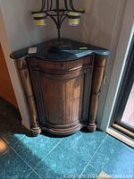 Full view of dark stone top wooden corner cabinet showing its curved shape, door, and column details.