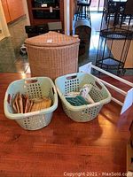View of corner wicker hamper with two plastic laundry baskets on a wooden floor; the baskets contain various hangers made of wood, plastic, and metal.