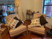 Two beige patterned arm chairs with wooden legs, each with two decorative pillows: one with an oval pattern in neutral tones and one solid dark brown pillow.