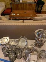 View of all silver plated serving ware including soup tureen, trays, butter dish, and mugs on a table with a couch and furniture in the background.