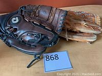 Two softball gloves side by side on a wooden surface, dark brown Demarini glove partially on top of tan Sasaki glove.
