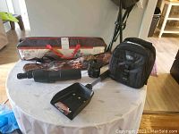 Photo showing all camping gear items on a round white table including the tent bag, monocular, shovel, air pump, tote bags, and stool in the background.