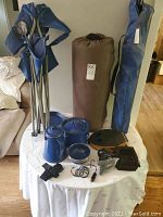 Photo showing two blue foldable camping chairs, brown rolled air mattress, blue metal camping dishes and small accessories on a white table.