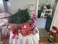 Artificial Christmas tree segments on top of a box with red bow, stockings, and clear container of assorted pink and purple ornaments on a white cloth-covered table