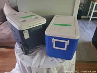 Two large Coleman coolers placed side-by-side on a round table. One cooler is dark blue with wheels and white lid; the other is blue with white lid and no wheels.