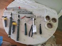 Photo displaying four paint rollers, a caulking gun, three rolls of painter's tape, a white tarp underneath, and a pair of pliers on a round table.