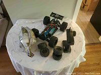 Remote-control car with large rubber tires, additional spare tires, black remote controller, and carry bag on round white table.