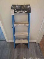 Full front view of blue 4-step Werner aluminum ladder with black top tray and aluminum steps placed on wooden floor against wall.