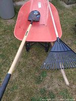 Full view of red-orange leading contractor wheelbarrow with black metal frame, wooden supports, and wheel, shown with yard rake and shovel inside wheelbarrow