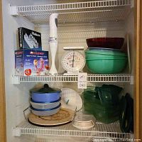 Shelf view showing Tupperware bowls, kitchen scale, immersion blender, apple peeler box, pizza stones, casserole containers and other kitchenware.
